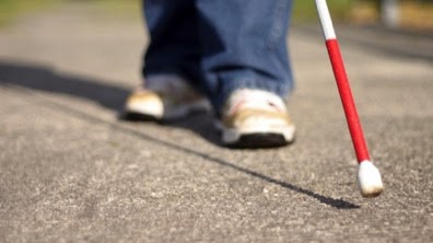 Photo: A close-up of the bottom of a long cane with cane tip positioned near a asphalt walkway, with the cane users feet walking in the background.