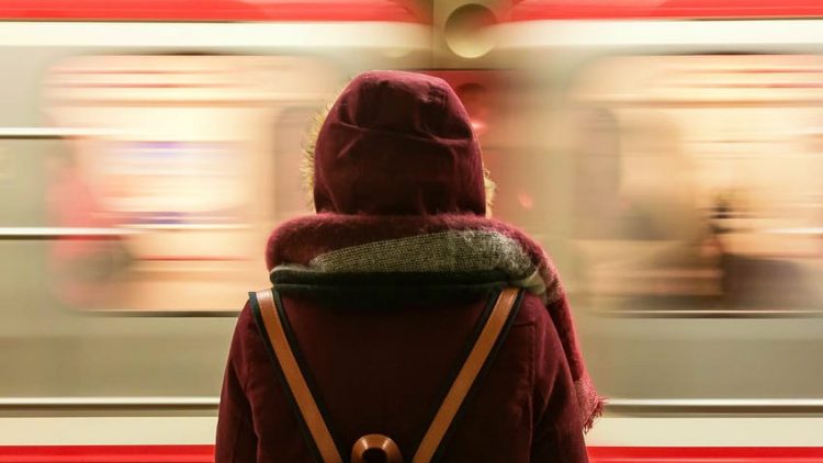 Photo: An individaul wearing a hooded coat, scarf, and backpack stamds with their back to the camera. A light rail train car appears to move quicly in front of them, as if they are waiting on a platform.